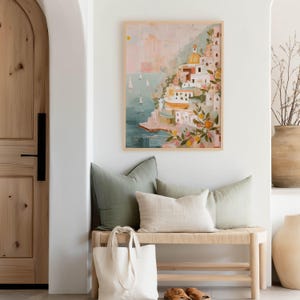 K&ouml;nnte beinhalten: Ein gerahmter Kunstdruck zeigt ein italienisches K&uuml;stendorf mit wei&szlig;en Geb&auml;uden, einer gelben Kuppel und Segelbooten auf dem Wasser. Das Kunstwerk befindet sich &uuml;ber einer Bank mit gr&uuml;nen und beigen Kissen, einer Tragetasche und einer gro&szlig;en Vase.