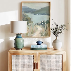 May include: A wooden cabinet with woven doors holds a green and blue ceramic lamp with a white lampshade. A framed landscape painting hangs above the cabinet. A small blue decorative bowl and a white vase with greenery are also on the cabinet.