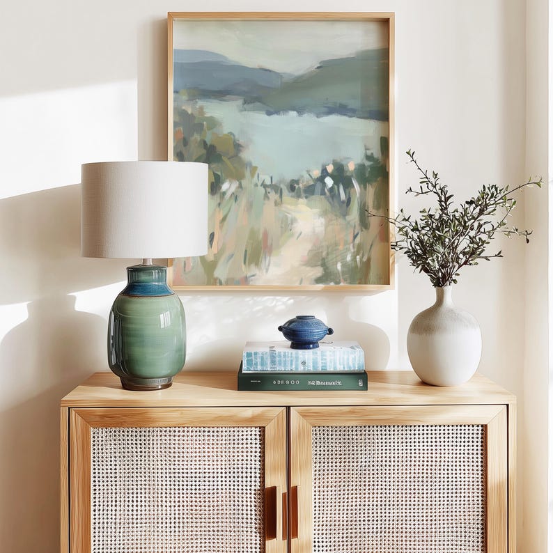 May include: A wooden cabinet with woven doors holds a green and blue ceramic lamp with a white lampshade. A framed landscape painting hangs above the cabinet. A small blue decorative bowl and a white vase with greenery are also on the cabinet.