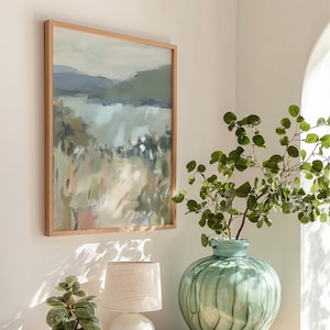 May include: A framed landscape painting with a light wood frame hangs on a white wall. The artwork depicts a serene scene with muted greens, blues, and beige tones, suggesting a natural landscape. A green vase with leafy branches sits nearby.