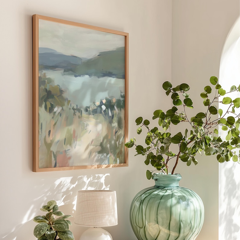 May include: A framed landscape painting with a light wood frame hangs on a white wall. The artwork depicts a serene scene with muted greens, blues, and beige tones, suggesting a natural landscape. A green vase with leafy branches sits nearby.