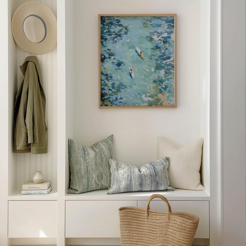May include: A coastal-themed interior scene featuring a framed artwork of surfers in the ocean, decorative pillows, a woven tote bag, and a straw hat. The color palette includes shades of blue, green, and beige, creating a serene atmosphere.
