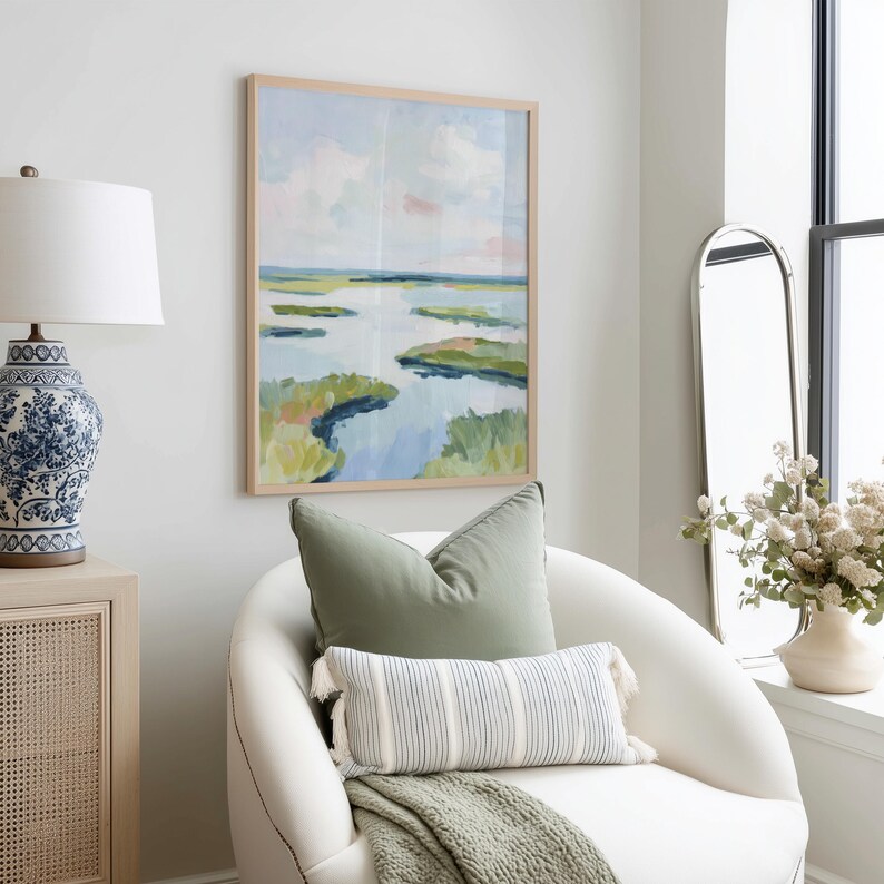 May include: A framed landscape painting with a light wood frame hangs above a white armchair. The painting depicts a marsh scene with blue water, green marsh grass, and a light blue sky. A green pillow and a striped pillow with tassels rest on the chair.