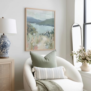 May include: A framed landscape painting with a light wood frame hangs above a white armchair. The painting depicts a serene scene with a lake, mountains, and fields of green, pink, and beige. A sage green pillow and a striped lumbar pillow with tassels rest on the chair.