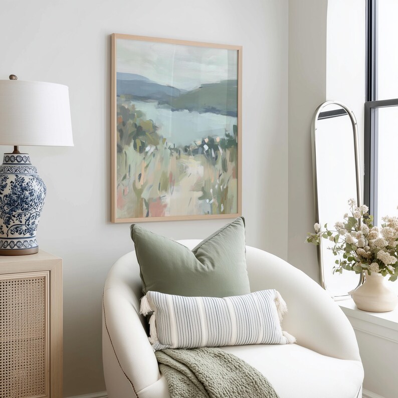May include: A framed landscape painting with a light wood frame hangs above a white armchair. The painting depicts a serene scene with a lake, mountains, and fields of green, pink, and beige. A sage green pillow and a striped lumbar pillow with tassels rest on the chair.