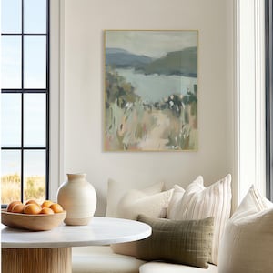 May include: An abstract landscape painting in a gold frame. The artwork depicts a path leading to a body of water, with muted tones of green, blue, and beige. The painting hangs above a round table with a wooden bowl of oranges.