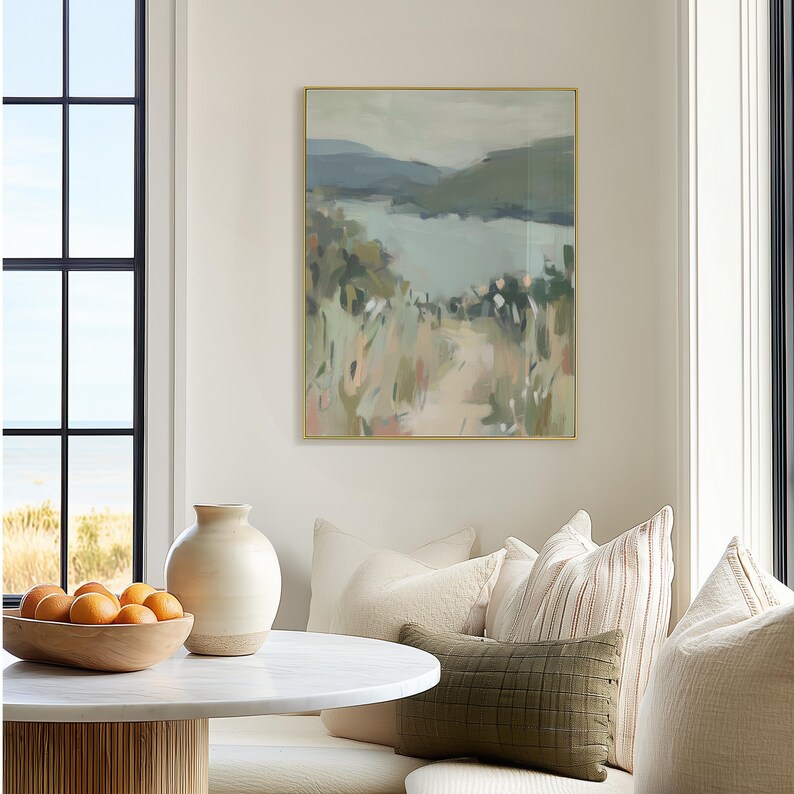 May include: An abstract landscape painting in a gold frame. The artwork depicts a path leading to a body of water, with muted tones of green, blue, and beige. The painting hangs above a round table with a wooden bowl of oranges.