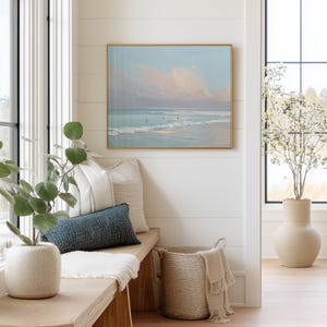 May include: A coastal-themed interior scene featuring a framed painting of a beach with a gold frame. A bench with pillows and a woven basket with a blanket are in the foreground. A large vase with a plant is in the background.