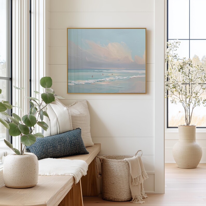 May include: A coastal-themed interior scene featuring a framed painting of a beach with a gold frame. A bench with pillows and a woven basket with a blanket are in the foreground. A large vase with a plant is in the background.