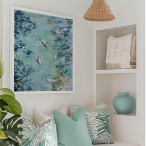 May include: A framed abstract art print depicts two people on surfboards in turquoise water. The artwork hangs above a bench with decorative pillows in shades of green and white. A light blue vase and a woven pendant light are also visible.