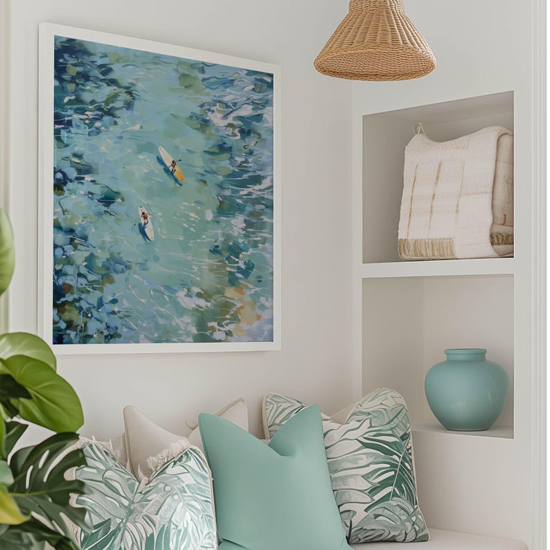 May include: A framed abstract art print depicts two people on surfboards in turquoise water. The artwork hangs above a bench with decorative pillows in shades of green and white. A light blue vase and a woven pendant light are also visible.