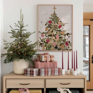 May include: A festive holiday scene featuring a small Christmas tree in a white pot, adorned with fairy lights. A framed abstract painting of a Christmas tree hangs above a wooden cabinet holding wrapped gifts and burgundy candles in white holders.