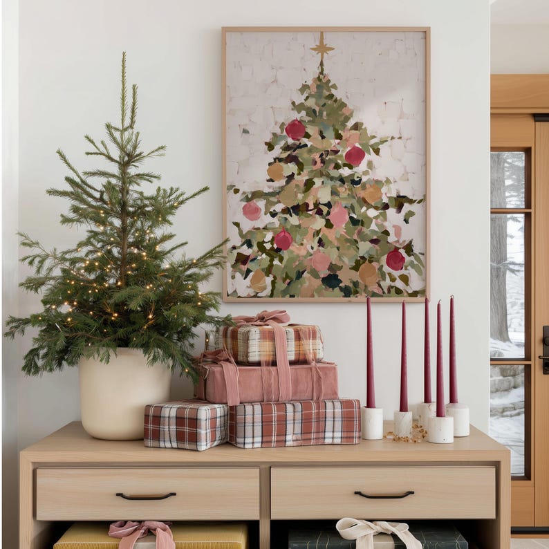 May include: A festive holiday scene featuring a small Christmas tree in a white pot, adorned with fairy lights. A framed abstract painting of a Christmas tree hangs above a wooden cabinet holding wrapped gifts and burgundy candles in white holders.