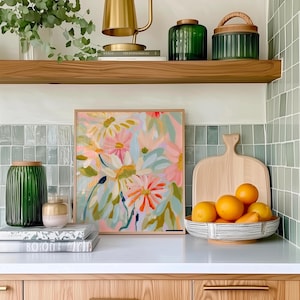 May include: A framed floral painting with pink, orange, and green flowers on a white background. The painting is hanging on a wall above a white countertop with a wooden cutting board and a bowl of oranges.