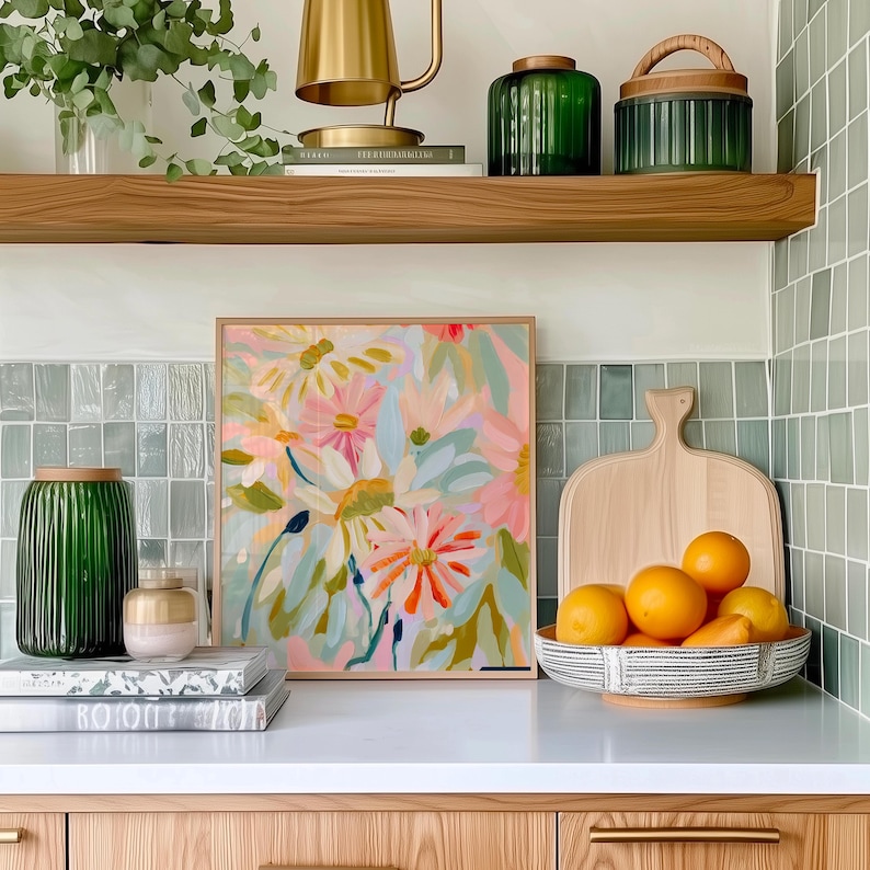 May include: A framed floral painting with pink, orange, and green flowers on a white background. The painting is hanging on a wall above a white countertop with a wooden cutting board and a bowl of oranges.