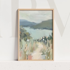 Op de afbeelding: Een ingelijst landschapsschilderij met een licht houten frame. Het kunstwerk toont een serene scène van een meer omgeven door bergen en groen. Het kleurenpalet omvat gedempte blauwe, groene en beige tinten, wat een kalmerende esthetiek creëert.