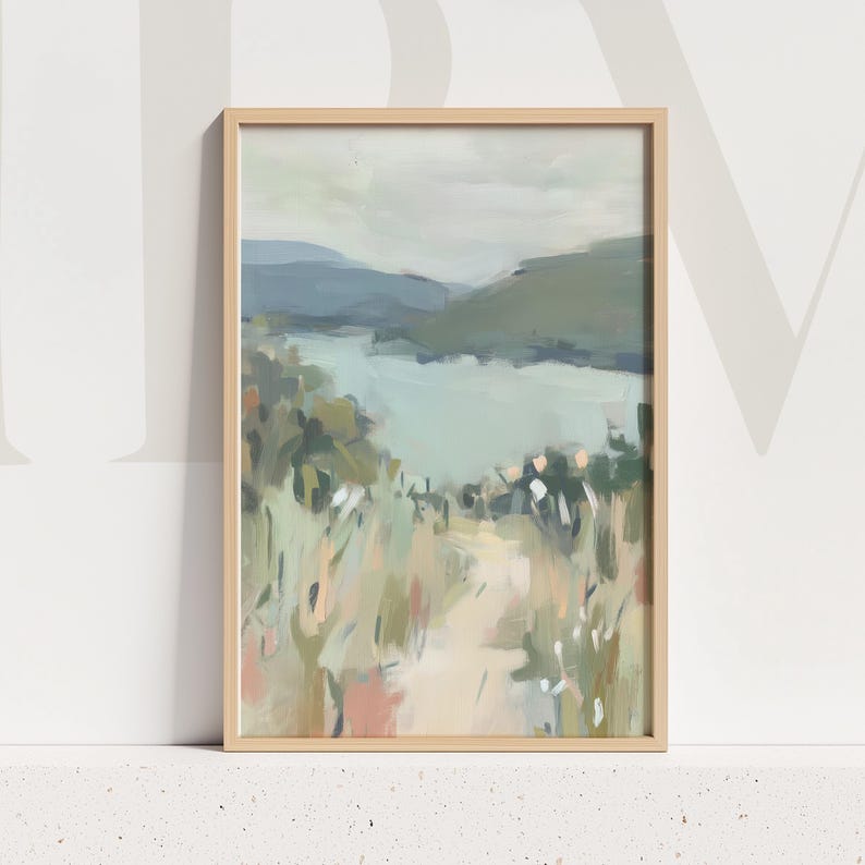 May include: A framed landscape painting with a light wood frame. The artwork depicts a serene scene of a lake surrounded by mountains and greenery. The color palette includes muted blues, greens, and beige tones, creating a calming aesthetic.