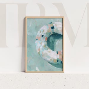 Puede incluir: Lámina artística enmarcada que presenta una pintura abstracta de un salvavidas. El salvavidas es blanco con parches rosas, azules y verdes. El fondo es de color verde azulado suave y el marco es de madera clara.