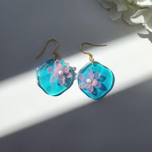 Peut inclure: Une paire de boucles d'oreilles en résine bleu turquoise avec une fleur rose et un accent de perle.