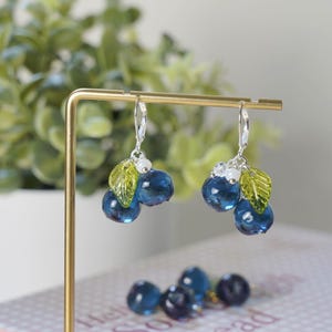 May include: A pair of dangle earrings featuring clusters of blue glass beads resembling blueberries. Each earring has a small green leaf and white bead accents. The earrings are displayed on a gold-colored stand against a blurred background.
