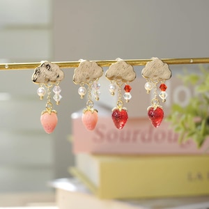Puede incluir: Cuatro pares de pendientes colgantes. Cada pendiente presenta una parte superior dorada en forma de nube, una cadena de perlas y cristales, y un dije de fresa. Dos pares tienen fresas rosas y dos tienen fresas rojas.
