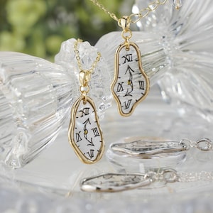 Puede incluir: Collares dorados con un diseño de colgante de reloj derritiéndose. Los colgantes presentan un fondo blanco con números romanos negros y manecillas de reloj. Los collares se exhiben sobre una superficie clara y reflectante.