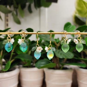Op de afbeelding: Een paar groene en blauwe citroenoorbellen met witte bloemaccenten. De oorbellen hangen aan een gouden standaard.