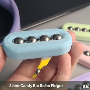 May include: A light blue oblong fidget toy featuring four embedded metallic silver spheres in a row. The device is labelled Silent Candy Bar Roller Fidget and ItJustClicks3D. Additional fidget toys in pastel yellow, pink, green, and purple are visible.