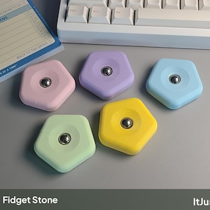 May include: Five pentagonal fidget stones in pastel pink, lavender, light blue, mint green, and yellow. Each matte-finished stone features a centred circular indentation containing a smooth, polished metal ball bearing for tactile sensory input.