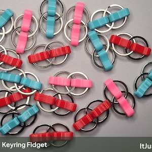 May include: A collection of fidget keyrings, each consisting of two metal split rings connected by a small, textured plastic bar in red, pink, or light blue. The text Keyring Fidget and ItJustClicks3D is printed at the bottom of the image.