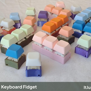 May include: A collection of keyboard fidget toys featuring mechanical switches and colorful keycaps in shades of white, blue, purple, orange, pink, and light green, mounted on various plastic bases. The text Keyboard Fidget and ItJustClicks3D appears.