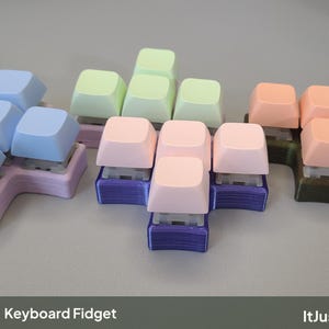 Puede incluir: Tres juguetes fidget de teclado con interruptores mecánicos y teclas en azul pastel, verde claro y melocotón. Cada base impresa en 3D es de color lavanda, verde claro o marrón oscuro. El texto Keyboard Fidget e ItJustClicks3D es visible.
