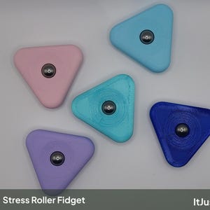 May include: Five triangular fidget rollers in pastel pink, light blue, teal, lavender, and dark blue. Each device features a central metallic ball bearing. The text Stress Roller Fidget and ItJustClicks3D is printed at the bottom of the image.