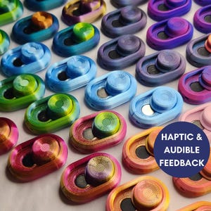 May include: A collection of colourful, oval-shaped, 3D printed objects with a round button in the centre. The objects are arranged in rows and have a variety of colours, including blue, green, red, purple, and pink. The text "Haptic & Audible Feedback" is visible in the bottom right corner of the image.