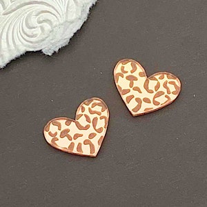 May include: Two heart-shaped wooden earrings with a leopard print design. The earrings are light brown with dark brown spots. A penny is next to the earrings for size comparison. The background is dark gray with a white textured paper.