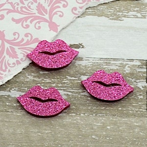 Op de afbeelding: Drie roze glitter lippen versieringen zijn gerangschikt op een licht houten oppervlak, in de buurt van een gescheurd stuk papier met een roze bloemenpatroon. Een cent is ook aanwezig, als maatreferentie.
