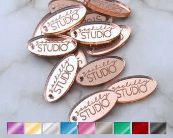 May include: A pile of rose gold oval-shaped acrylic tags with the text "saelilly STUDIO" engraved on each tag.
