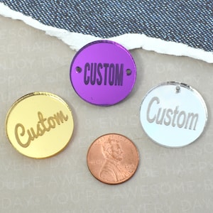 以下が含まれることがあります： 「Custom」という単語が異なるスタイルと色で書かれた、丸い鏡面アクリルタグ3つ。タグは金色、銀色、紫色です。