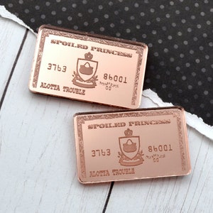 Op de afbeelding: Twee rechthoekige rozegouden pins met de tekst "SPOILED PRINCESS" en "ALOTTA TROUBLE". Elke pin heeft een schildontwerp met een kroon en een handtas. Een koperen penny ligt naast de pins.