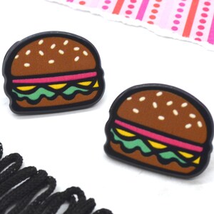 May include: Two small, cartoon-style hamburger earrings. The earrings are brown, yellow, green, and pink.