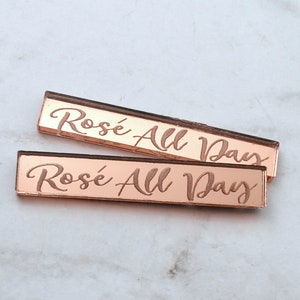 May include: Two rose gold acrylic signs with the text "Rosé All Day" in a cursive font. The signs are about the size of a US penny.