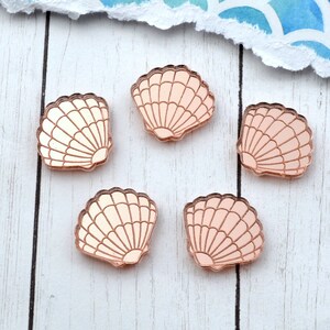 Puede incluir: Cinco formas de concha de acrílico de color oro rosa sobre una superficie de madera blanca. Se incluye una moneda de un centavo como referencia de tamaño.