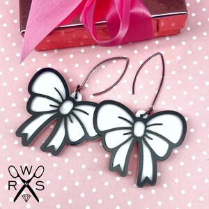 May include: A pair of white bow earrings with black outlines. The earrings are on a pink background with white polka dots.