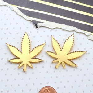 May include: Two gold-colored, cannabis leaf-shaped cutouts. The leaves have detailed edges and are placed on a white surface with small gray polka dots. A penny is next to the leaves for size comparison. The background includes torn paper with black and gold stripes.