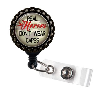 以下が含まれることがあります： 「Real Heroes Don't Wear Capes」と書かれた迷彩柄の丸い金属製チャームが付いた、黒いリトラクタブルバッジホルダーです。