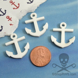 Può includere: Tre ciondoli a forma di ancora in legno bianco, ognuno delle dimensioni di una moneta da un centesimo di euro. Le ancore hanno un design semplice con un anello in cima.