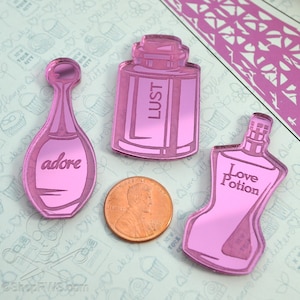 May include: Three pink acrylic perfume bottle shapes with the words "adore", "LUST", and "Love Potion". A penny is next to the shapes for size comparison. The background is a light blue and white pattern with the words "New York" and "love letters".
