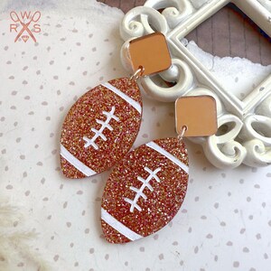 May include: A pair of glittery football-shaped earrings. The earrings have a copper-colored glitter base with white stripes and stitching details. The top of the earrings are square-shaped and copper-colored.