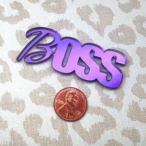 May include: Purple iridescent acrylic word "Boss" with a stylized font. The word is about the size of a US penny.