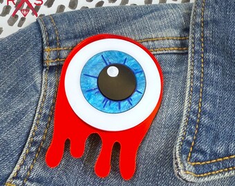 Pode incluir: Broche em forma de olho com íris azul, esclerótica branca e pupila preta, sobre um fundo vermelho gotejante. Preso a uma jaqueta jeans, apresenta um acessório artístico e arrojado.